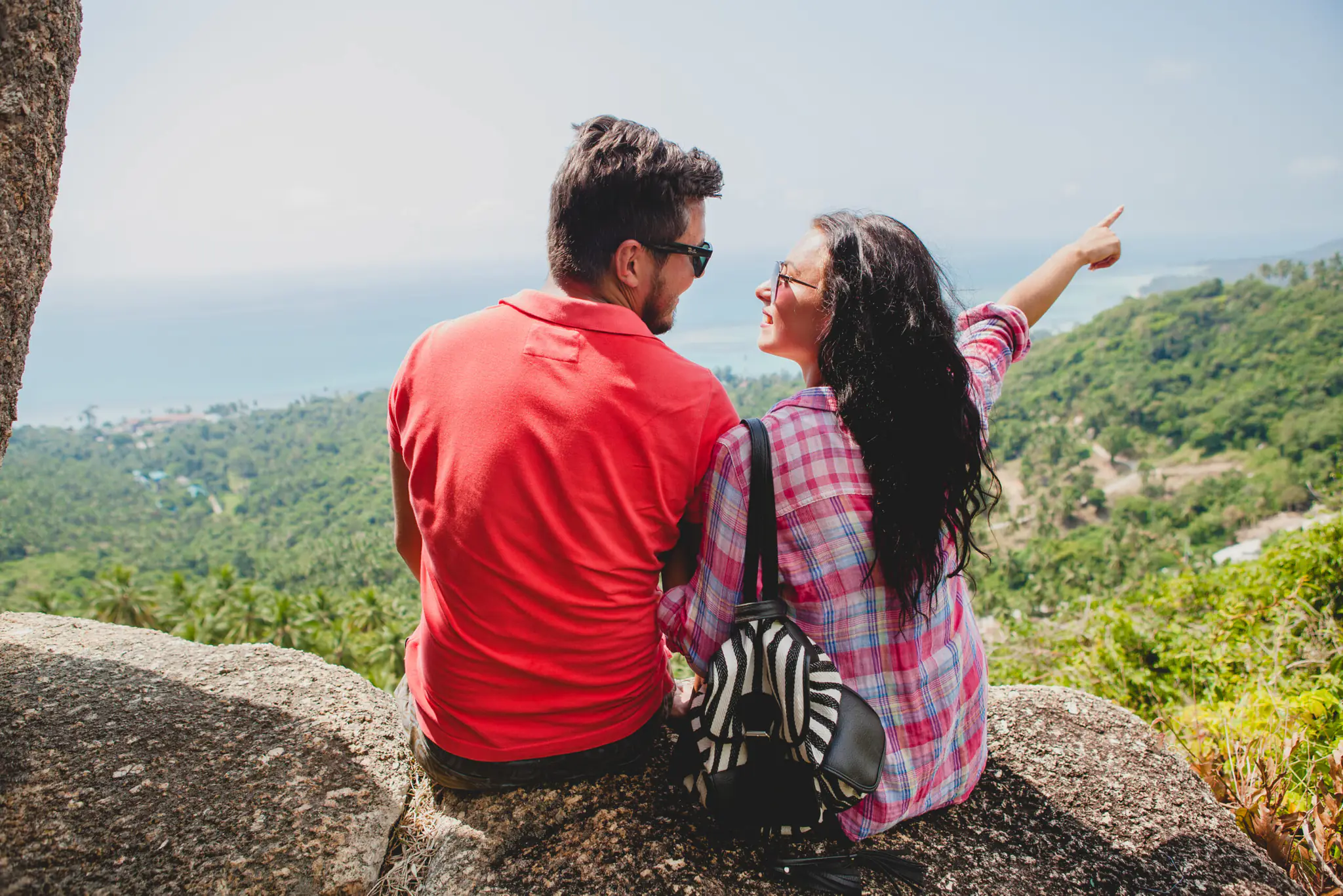 young happy hipster couple in love traveling around world, sightseeing, tropical romantic vacation in thailand, sitting on a rock, pointing finger, active tourism, summer